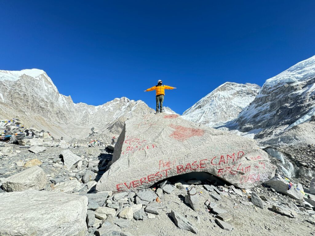 A mountaineer stands on a rock at Everest Base Camp, surrounded by snowy peaks.